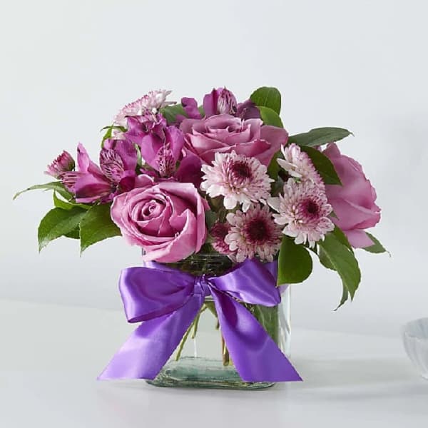 Pink roses and daisy-like flowers in a glass vase with a purple ribbon