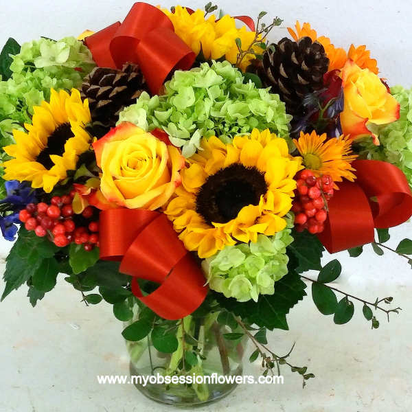 Bouquet of sunflowers, yellow roses, and green hydrangeas in a glass vase with red ribbon