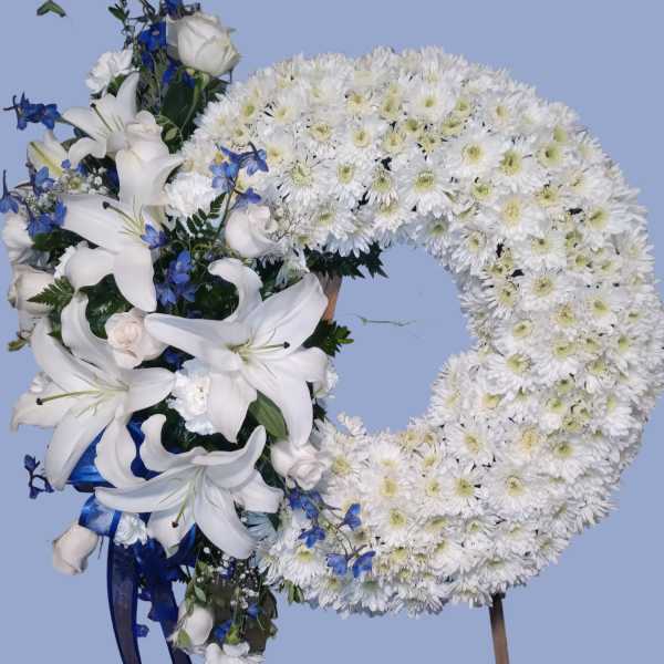 White floral wreath with lilies and blue accents on a stand