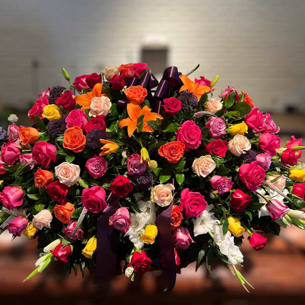 Large multicolored rose and lily casket spray with a purple ribbon bow arranged in an oval shape.