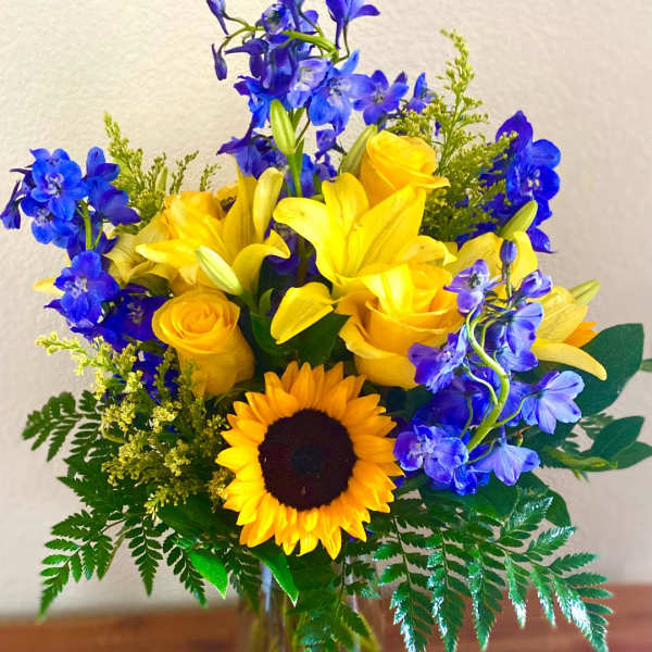 Yellow roses, lilies, and a sunflower in a glass vase with blue flowers