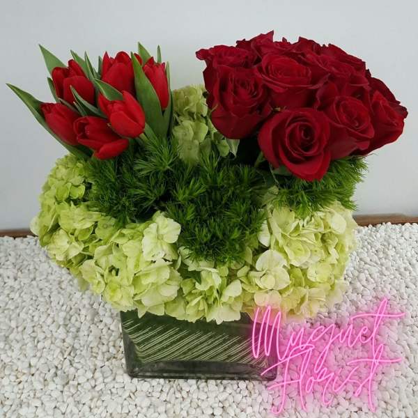 Red roses and tulips arranged with pale green hydrangeas in a square vase.