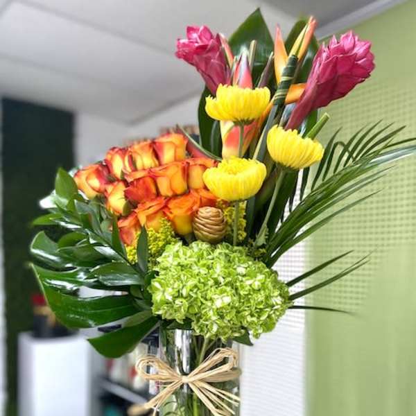 Tall tropical arrangement with orange roses, green hydrangea and yellow blooms in a glass vase with raffia bow