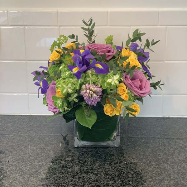 Mixed bouquet of pink roses, yellow spray roses, and purple irises in a square glass vase