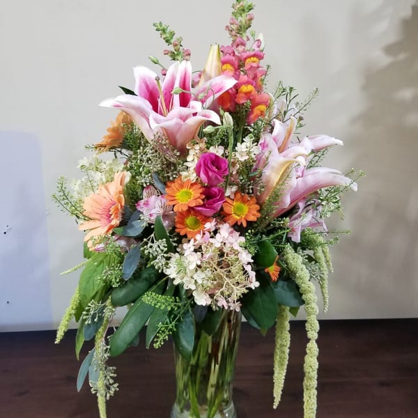 Tall mixed arrangement of pink lilies, orange daisies, and assorted flowers in a clear glass vase