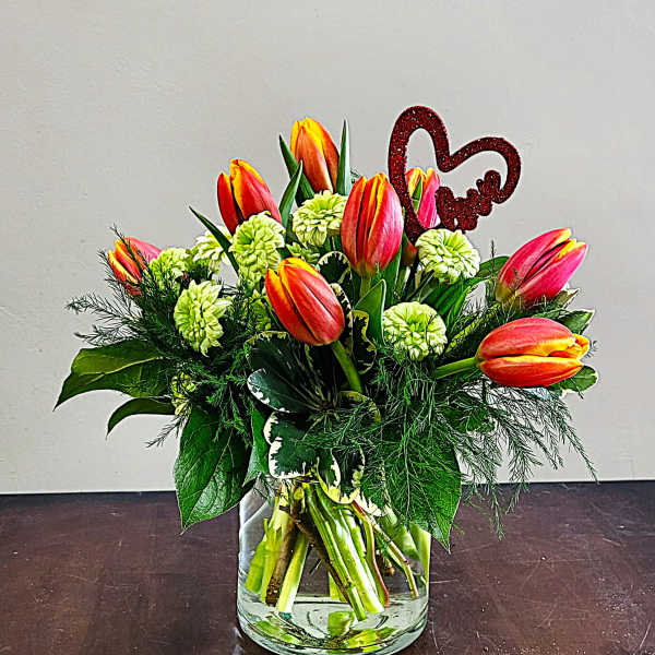 Short vase arrangement of orange tulips and green button flowers with a red glitter heart pick
