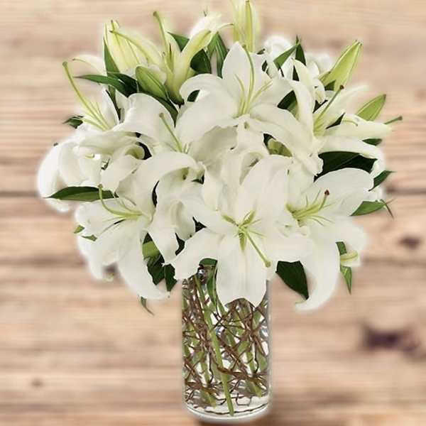 Tall arrangement of white lilies in a clear glass cylinder vase with visible stems.
