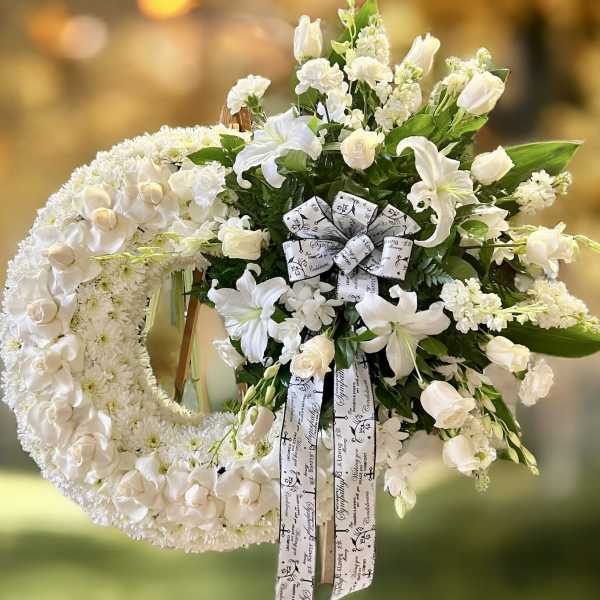 Large white floral wreath with side spray of lilies and roses on a standing easel with a printed ribbon