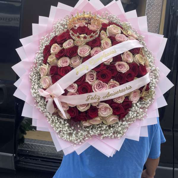 Large round bouquet of red and blush roses with pink wrapping and a crown topper