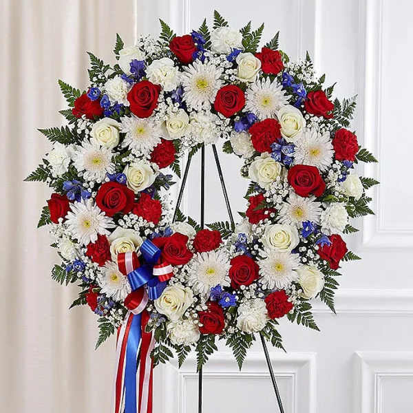 Patriotic standing wreath of red, white, and blue flowers with a striped ribbon on an easel.