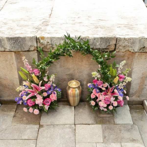 Two floral sprays flank a gold urn on a stone surface.