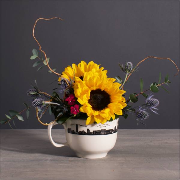 Compact arrangement of sunflowers, purple thistles, and red blooms in a white skyline mug