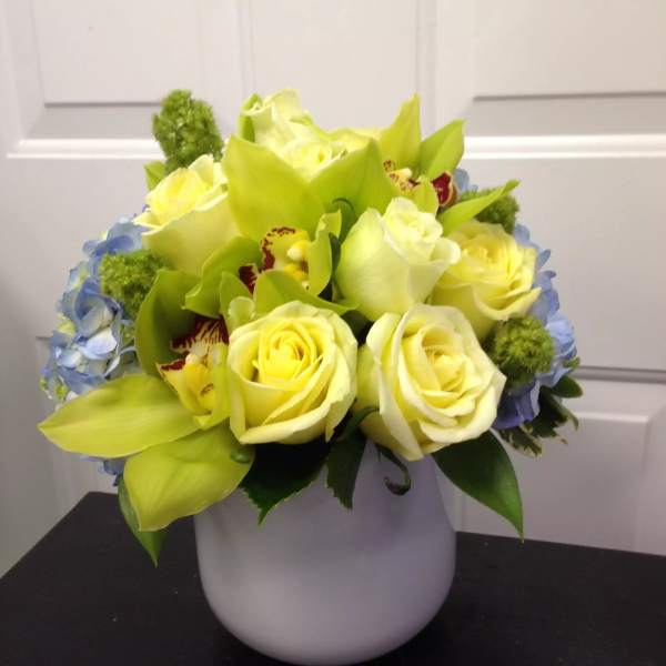 Yellow roses and green orchids in a white vase with blue hydrangea accents