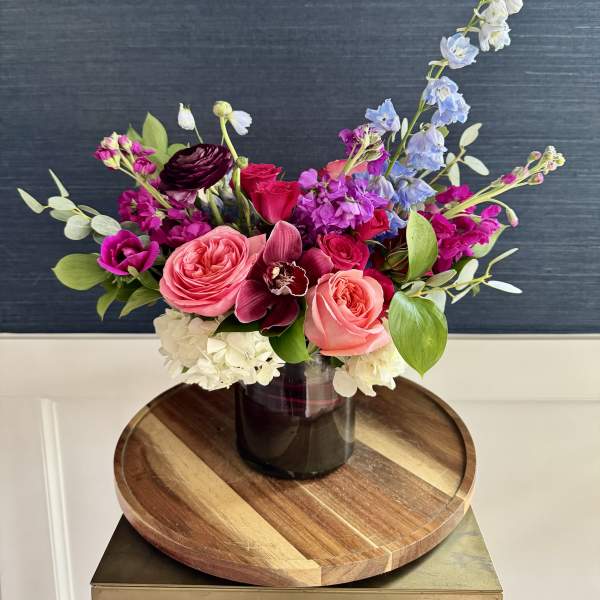 Mixed arrangement of pink roses, white hydrangeas, and purple blooms in a dark glass vase on a wooden tray