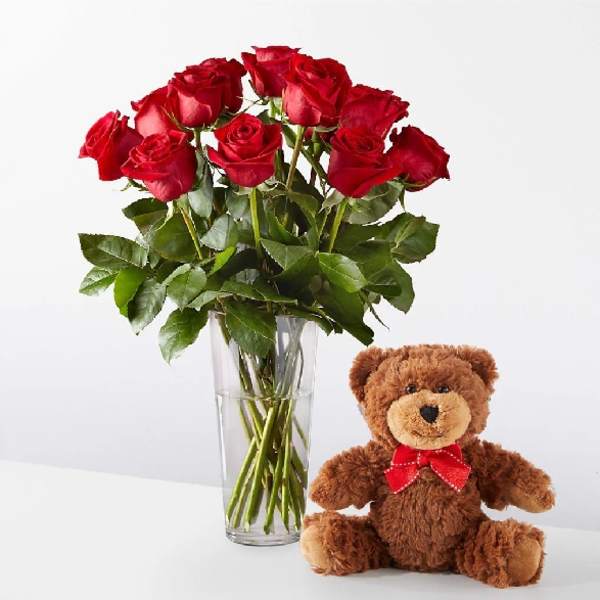 Red roses in a clear vase beside a teddy bear with a red bow