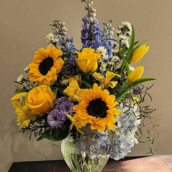 Arrangement of yellow sunflowers, roses, and blue hydrangeas in a clear glass vase