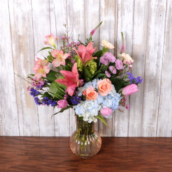 Tall mixed bouquet of pink lilies, roses, tulips, blue hydrangeas and purple blooms in a clear ribbed glass vase