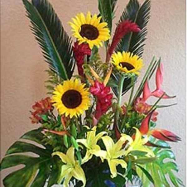 Tall tropical arrangement with yellow sunflowers, lilies, and red-orange flowers in a dark vase