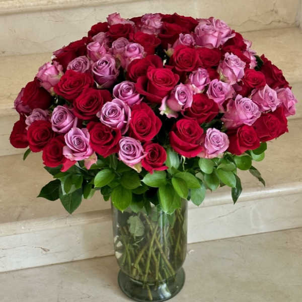 Large arrangement of red and lavender roses in a clear glass vase with a lush green collar