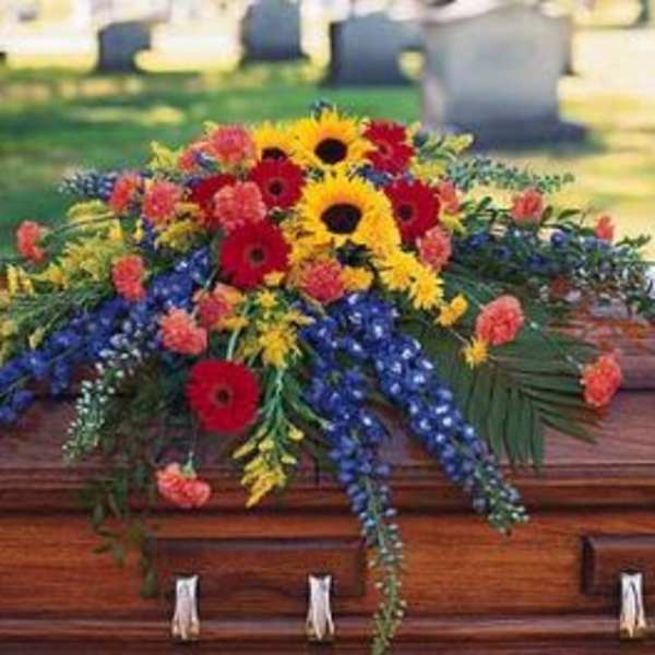 Bright mixed casket spray with sunflowers, red gerbera daisies, and blue flowers on a wooden casket