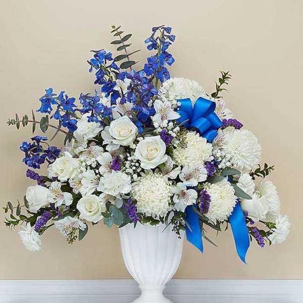 Tall white and blue sympathy flower arrangement in a white pedestal vase with a blue bow