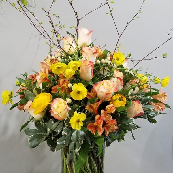 Tall vase arrangement with peach roses, yellow ranunculus, and orange alstroemeria accented by airy branches.