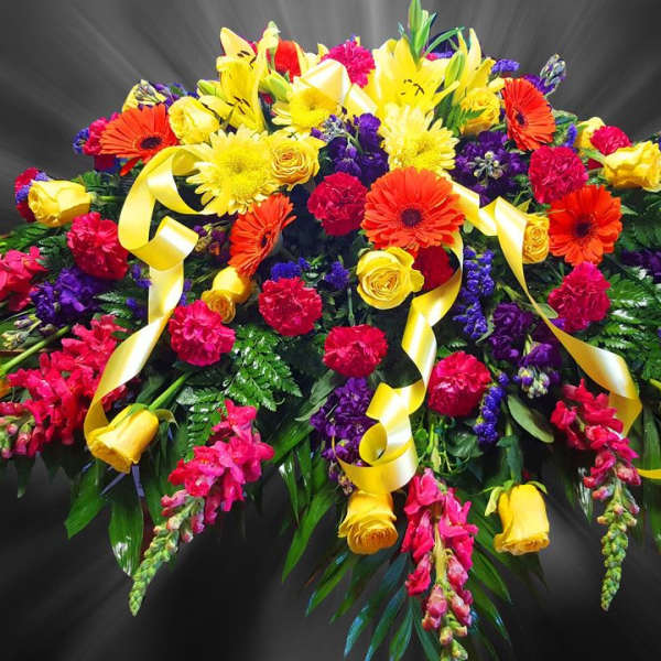 Bright mixed casket spray with yellow roses, orange gerberas, red carnations, purple blooms and yellow ribbons.