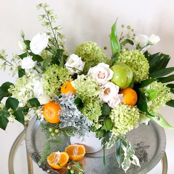 White and green floral arrangement with oranges and a green apple in a white vase