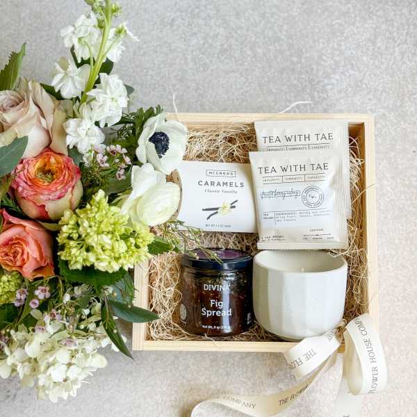 Floral gift box with roses and white blooms beside tea, caramels, fig spread, and a mug