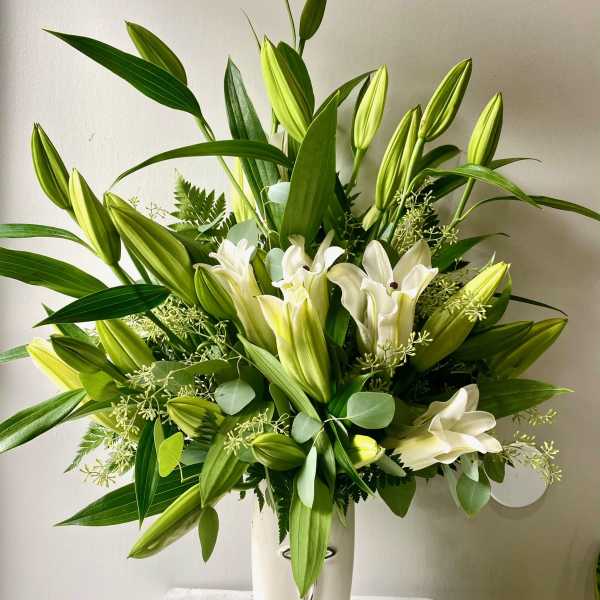 White lilies in a tall white vase with lush green foliage