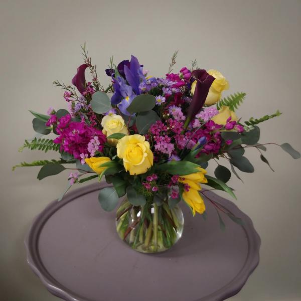 Mixed bouquet of yellow roses, purple flowers, and dark calla lilies in a glass vase