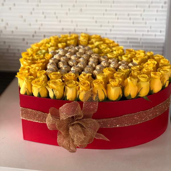 Yellow roses arranged in a red hat box with gold ribbon and chocolates in the center