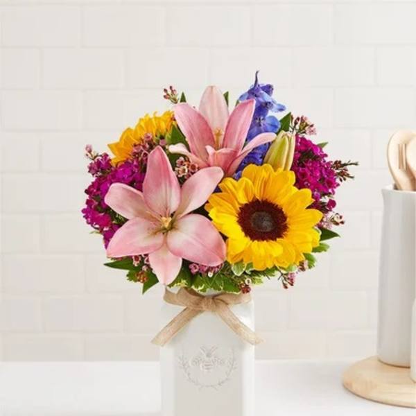 Colorful bouquet of pink lilies, a yellow sunflower, and mixed blooms in a white ceramic vase with a bow.