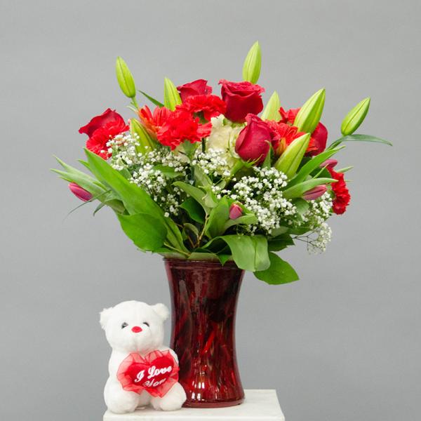 Red bouquet in a glass vase with a small teddy bear holding a heart