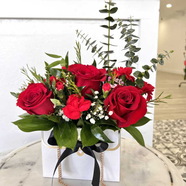 Red roses arranged in a white gift box with greenery and a black ribbon