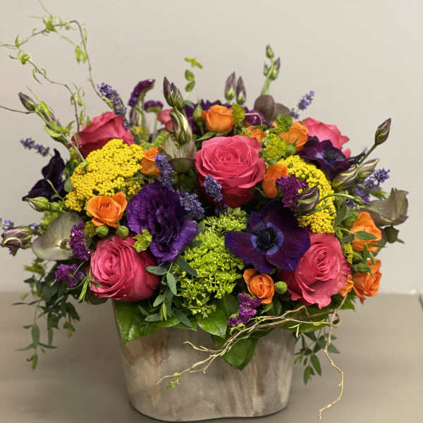 Vibrant arrangement of pink roses, purple blooms, orange flowers and green accents in a rustic ceramic container.
