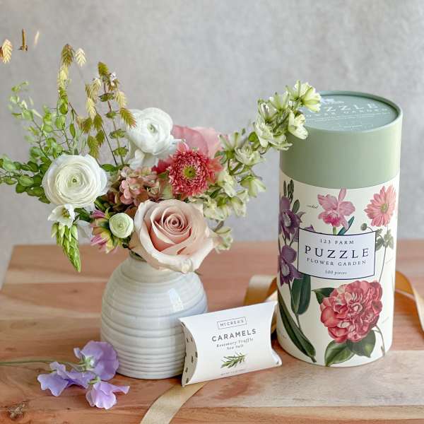 Mixed flower arrangement in a white vase beside a floral puzzle tin and caramel box