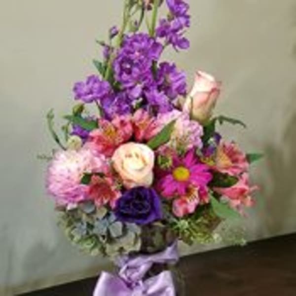 Tall mixed bouquet of purple and pink flowers with roses and daisies in a glass vase tied with a lavender bow