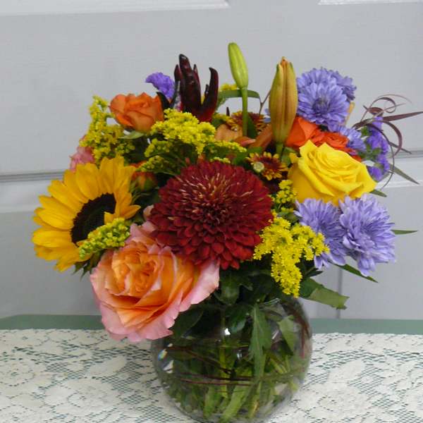 Mixed bouquet in a glass vase with roses, sunflowers, lilies, and purple blooms
