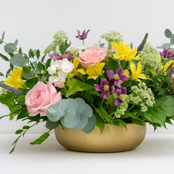 Mixed bouquet of pink roses, yellow flowers, and purple blooms in a gold bowl