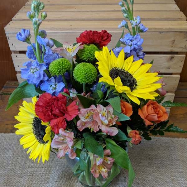Mixed bouquet with sunflowers, roses, carnations, and blue flowers in a glass vase