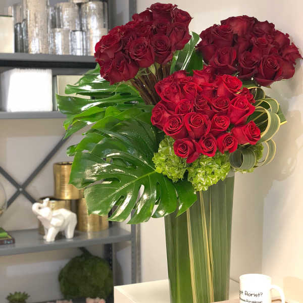 Tall arrangement of clustered red roses with large tropical green leaves in a clear glass vase