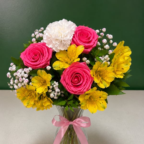 Pink roses, white carnation, and yellow alstroemeria in a glass vase with a pink ribbon