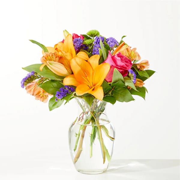 Bright mixed bouquet of lilies, roses, and purple filler flowers in a clear glass vase
