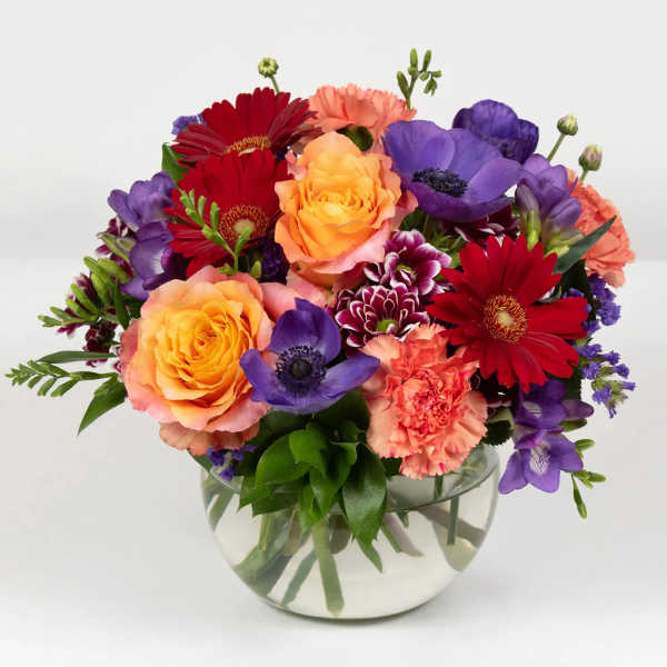 Mixed bouquet of roses, gerbera daisies, carnations, and purple blooms in a glass bowl vase
