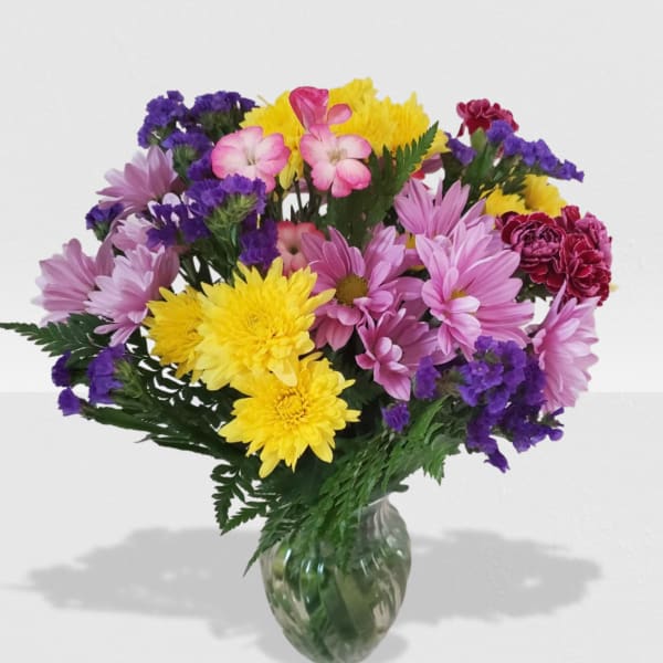 Vase of mixed pink daisies, yellow mums, purple filler flowers, and red carnations.