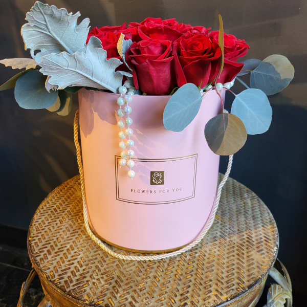 Red roses arranged in a pink hatbox with pearl strands and blue-gray foliage.