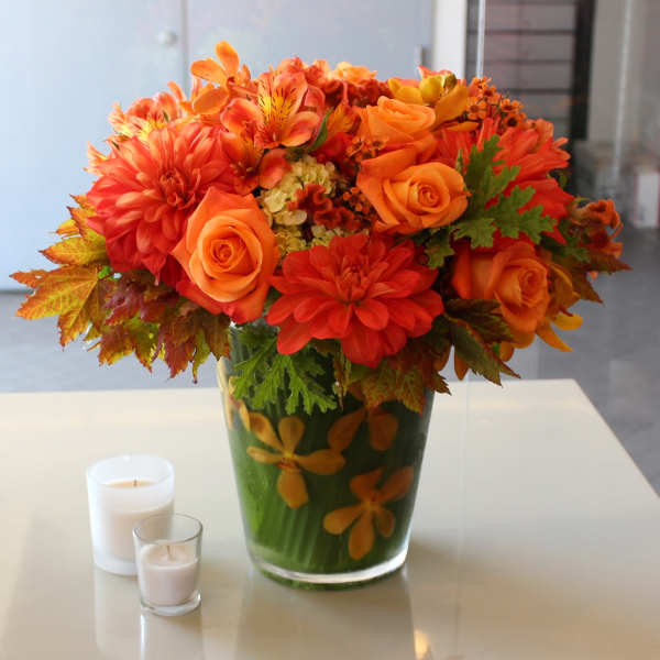 Orange floral arrangement in a glass vase with two candles nearby