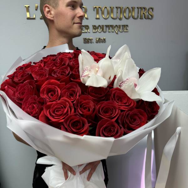 Large bouquet of red roses with white orchids wrapped in white paper, held by a person indoors