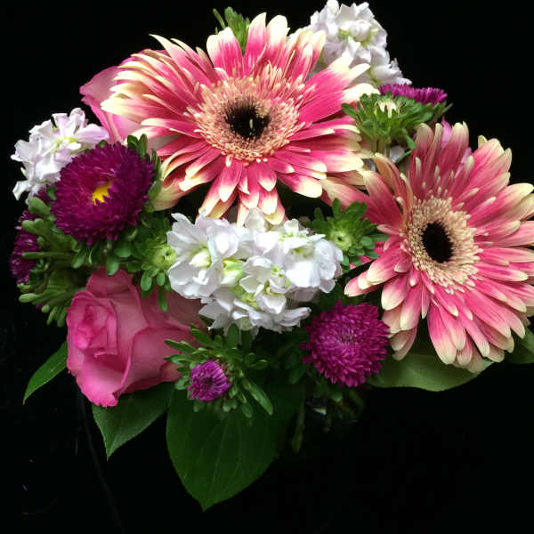 Pink gerbera daisies, roses, and white blossoms arranged in a small vase against a black background